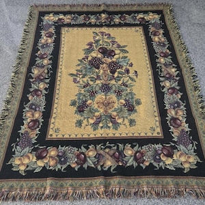 May include: A woven tapestry throw blanket with a black background and a central design of a fruit-bearing tree with a gold background. The border is a repeating pattern of fruit and leaves.