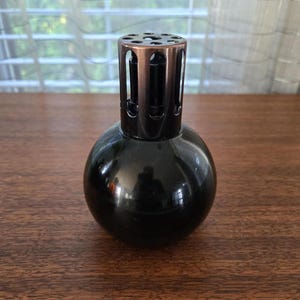 May include: A spherical black glass fragrance lamp featuring a metallic bronze-toned vented cap with circular cutouts at the top. The lamp has a smooth, reflective surface and a cylindrical neck that connects to the rounded base of the vessel.
