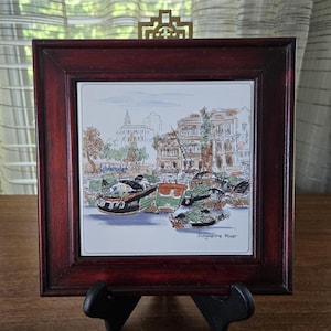 May include: A framed ceramic tile featuring a waterfront scene with boats and buildings. The tile is set in a dark wood frame with a decorative brass accent. The words "Singapore River" are written on the tile.