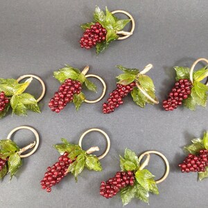 Op de afbeelding: Set van acht decoratieve servetringen. Elke ring heeft een tros rode druiven met groene bladeren en een goudkleurige ring. De druiven hebben een glanzende afwerking en de bladeren zijn doorschijnend.