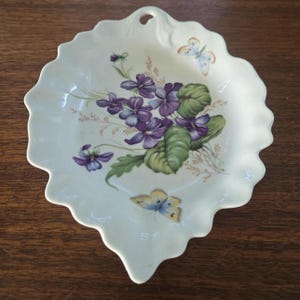 May include: A leaf-shaped, off-white ceramic dish with a scalloped edge. The dish is decorated with a cluster of purple violets, green leaves, and two butterflies. The dish has a small hole at the top.