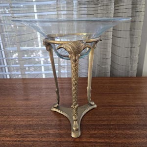 Puede incluir: Un cuenco decorativo de vidrio con un borde ancho, sostenido por un soporte de metal color latón. El soporte presenta un diseño de cabeza de carnero y detalles ornamentados. El cuenco es transparente y el soporte tiene una base de tres patas.