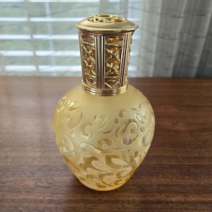 May include: A frosted yellow glass fragrance lamp with a bulbous body featuring an intricate, raised swirling pattern. The lamp is topped with a metallic gold-toned vented cap that has a decorative lattice design around its cylindrical neck.