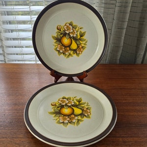以下が含まれることがあります： 茶色の縁取りが付いた白いセラミック製のディナー皿2枚。皿には、黄緑白のフラワーデザインが施され、洋梨と茶色の背景が描かれています。
