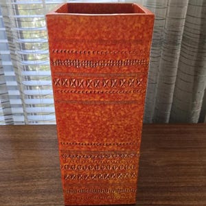Op de afbeelding: Een hoge, rechthoekige oranje vaas met een textuuroppervlak. De vaas heeft een herhalend patroon van horizontale lijnen, stippen en kruisvormige ontwerpen. De vaas is gemaakt van keramiek of een vergelijkbaar materiaal. Ongeveer 30 cm hoog.