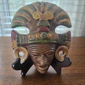 Könnte beinhalten: Eine braune, geschnitzte Maske mit detailliertem Gesicht, verziert mit grünen, goldenen und roten Akzenten. Die Maske wird auf einem schwarzen Ständer präsentiert, der ihr kompliziertes Design und ihre kulturelle Bedeutung hervorhebt. Die Maske hat ein verwittertes Aussehen.
