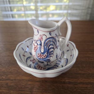 May include: A white ceramic jug and bowl set. The jug features a handle and a blue and red rooster design. The bowl has a scalloped edge and a matching floral pattern. The set is likely vintage or antique, with a traditional aesthetic.