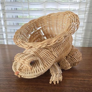 May include: A woven wicker frog-shaped basket with a wide, open mouth. The basket is made of natural, light brown wicker and has a slightly textured surface.