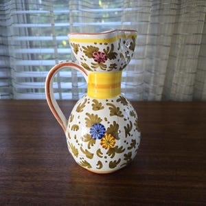 Op de afbeelding: Decoratieve keramische kan met een bolle basis en een uitlopende bovenkant. De kan is wit met een bloemmotief in bruin, geel, roze en blauw. Het heeft een gele band in het midden en een bruine handgreep.