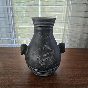 May include: A dark grey ceramic vase with a rounded body and a narrow neck. The vase has embossed figures of a person and an animal. It has two small handles and is placed on a wooden surface.