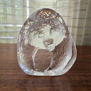 May include: A clear glass bear sculpture with a textured finish. The bear is shown in a seated pose, with its head gently lowered. The sculpture has an oval shape and is placed on a wooden surface.