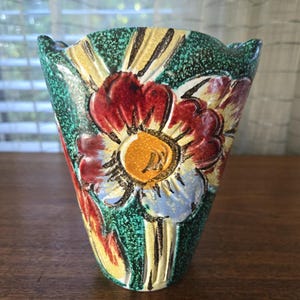 May include: A ceramic vase with a floral design. The vase features a green speckled background with a large flower in the center, showcasing red, yellow, and blue petals. The vase has a unique, scalloped rim and is set on a wooden surface.