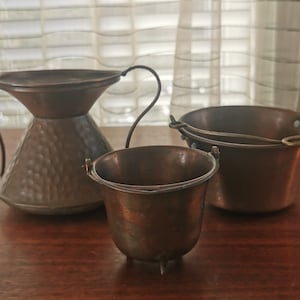 May include: Three copper pots of varying sizes. The largest pot has a handle and is slightly wider at the top. The smallest pot has three legs and a handle. The middle pot has a handle and is slightly wider at the top.