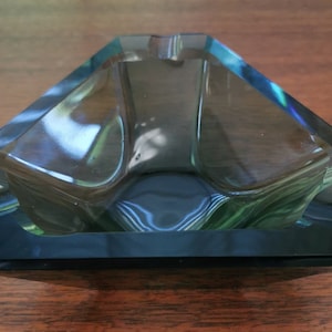 May include: A dark blue glass ashtray with a geometric, triangular shape. The ashtray has a deep well for holding cigarette butts.