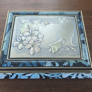 Puede incluir: Una caja de joyería rectangular con una tapa plateada con un diseño floral y un marco con estampado azul y negro. La caja tiene un borde dorado y un patrón floral a juego en la base, creando una estética vintage.