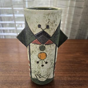 May include: A decorative ceramic vase with a unique geometric design. The vase features a cream-coloured base with dark green, black, orange, and yellow accents. The design includes geometric shapes and small circular details. The vase is cylindrical in shape.