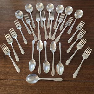 Pode incluir: Uma variedade de talheres de prata antigos, incluindo garfos e colheres, dispostos sobre uma superfície de madeira. Os talheres têm um acabamento prateado brilhante e detalhes ornamentais nas alças. A disposição sugere uma mesa formal.