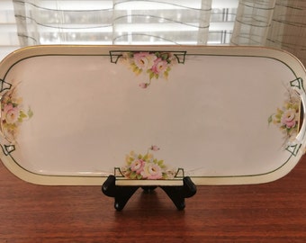 May include: An elongated, rectangular porcelain serving tray with rounded corners and gold-trimmed edges. The white surface features floral clusters of pink roses and green leaves at the center of each side, accented by thin, dark green geometric lines.