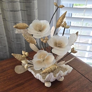 May include: A decorative arrangement featuring three white shell flowers with gold centres, accented by gold-painted spiral seashells and thin dark stems. The floral display is set within a large, white, fluted clam shell filled with light-coloured sand.