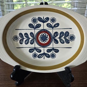 以下が含まれることがあります： 茶色の縁取りと青と白の花柄が施された白い楕円形のサービングプレート。デザインの中心には赤と白の円があります。