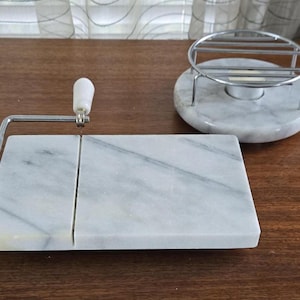 Puede incluir: Una tabla de queso de mármol blanco con un cortador de queso de alambre integrado. La tabla está dividida en dos secciones. Una base separada de mármol blanco con una rejilla de alambre cromado se encuentra junto a la tabla de queso.