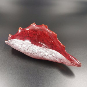 以下が含まれることがあります： 貝殻の形をした装飾ガラスボウル。ボウルは、白とピンクの斑点模様のインテリアと鮮やかな赤色を特徴としています。縁は質感のあるギザギザの縁です。ボウルは濃い灰色の表面にあります。