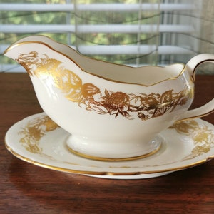 May include: A white porcelain gravy boat with a gold floral design and a matching saucer. The boat has a handle and a scalloped edge.
