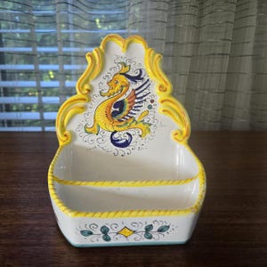May include: A white ceramic wall-mounted soap dish with yellow and gold ornate trim. The dish features a hand-painted mythical creature in yellow, blue, and orange. The base has a decorative floral design.