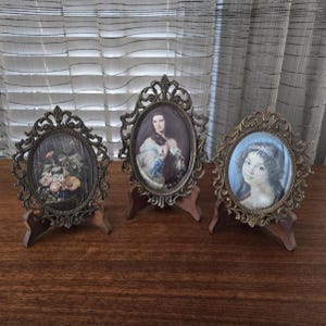 May include: Three antique oval picture frames with ornate bronze-coloured metalwork. Each frame holds a different portrait or still life image. The frames are displayed on small wooden stands, showcasing vintage decor.
