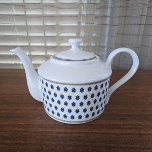 May include: White ceramic teapot with a lid and handle. The body is decorated with a repeating pattern of blue and yellow floral shapes. The teapot has a spout and a curved handle.