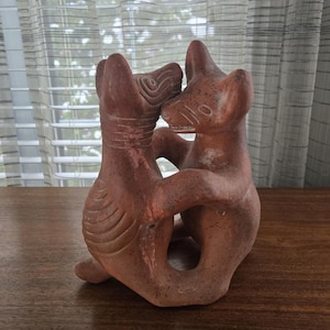 Pode incluir: Escultura em terracota castanho-avermelhada que retrata duas figuras de animais estilizadas a abraçar-se. As figuras têm formas simples e arredondadas. A escultura está colocada sobre uma superfície de madeira.