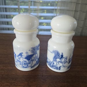 Könnte beinhalten: Zwei weiße Keramikgläser mit abgerundeten Deckeln. Jedes Glas hat ein blau-weißes Transferdruckdesign mit Szenen mit Menschen, Kutschen und Pferden. Die Gläser stehen auf einer Holzoberfläche.