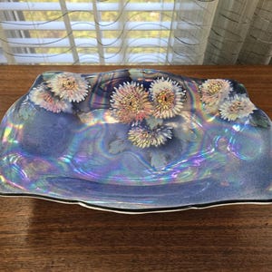 May include: Iridescent blue decorative dish with a floral design. The dish has a wavy shape and is decorated with white and pink chrysanthemum flowers. The rim is black with a white border. The dish is sitting on a wooden surface.
