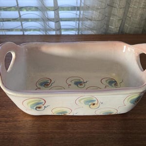 May include: Rectangular ceramic baking dish with handles. The dish is off-white with a pink rim and handles. It features a repeating pattern of blue, green, and yellow swirls. A decorative and functional kitchenware item.
