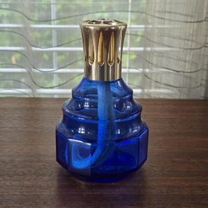 May include: A cobalt blue glass fragrance lamp with a faceted, hexagonal base and a tiered, rounded upper section. The lamp features a metallic gold-toned vented crown cap at the top and a thick, blue fabric wick extending into the interior.