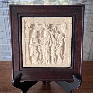 Pode incluir: Uma escultura em relevo emoldurada que retrata um grupo de figuras, provavelmente religiosas ou históricas, em bege claro. A escultura está emoldurada em uma moldura de madeira marrom escura e é exibida em um suporte preto.