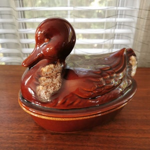 May include: A brown ceramic duck-shaped casserole dish with a lid. The dish has a glazed finish and a speckled pattern.