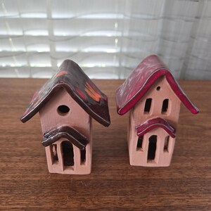 May include: Two ceramic miniature houses with pitched roofs. One has a brown roof with orange accents, the other a red roof. Both have rectangular windows and doors, and are painted in shades of beige and brown.