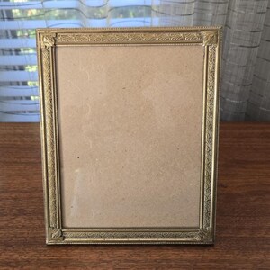 May include: A rectangular, antique gold-colored picture frame. The frame has a detailed, ornate design around the edges and corners. The frame is empty, with a light brown backing visible. The frame is sitting on a dark brown wooden surface.