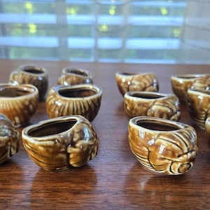 Könnte beinhalten: Eine Sammlung kleiner, brauner Keramikbehälter in Muschelform. Die Behälter haben eine glänzende Oberfläche und sind auf einer Holzoberfläche angeordnet. Das Muscheldesign ist detailliert und strukturiert.