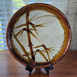 Puede incluir: Un plato redondo de cerámica con un borde marrón y un centro blanco. El plato presenta un diseño pintado a mano de ramas de bambú en marrón y dorado.