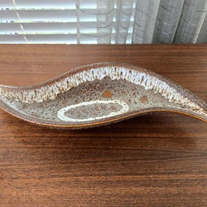 Puede incluir: Un plato de cerámica alargado y curvado con un esmalte moteado en tonos marrón, beige y crema. La superficie interior muestra un patrón texturizado y manchado con un distintivo acento ovalado blanco en el centro y un borde marrón oscuro.