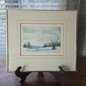 May include: A framed landscape print featuring a winter scene with a large bare tree in the foreground, a snow-covered field, and distant trees under a cloudy sky. The artwork is matted in cream and enclosed in a thin gold-toned metal frame.