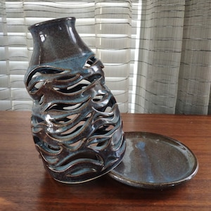 May include: A ceramic candle holder with a teal and brown glaze. The holder has a unique design with cutouts that resemble waves. It comes with a matching saucer.