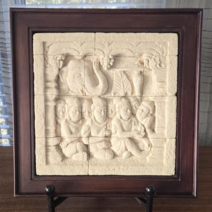Pode incluir: Escultura em relevo emoldurada, esbranquiçada, retratando figuras e um elefante. A escultura é montada em uma moldura de madeira marrom escura e exibida em um suporte de metal preto. O relevo parece ser esculpido em pedra.