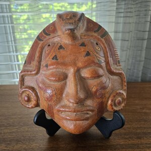 Könnte beinhalten: Eine terrakottafarbene Keramikmaske mit komplexen Schnitzereien, darunter eine zentrale Tierfigur an der Oberseite, geometrische Muster und kreisförmige Ohrverzierungen. Die Maske hat geschlossene Augen und einen neutralen Ausdruck auf einem schwarzen Ständer.