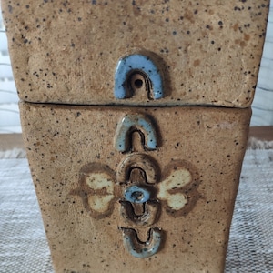 Puede incluir: Una caja de cerámica con un esmalte marrón moteado y detalles en azul y blanco. La caja tiene una tapa y un diseño decorativo en la parte delantera.