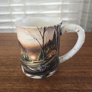 May include: Ceramic mug with a scenic camping design. The mug features a landscape with a sunset, trees, a tent, and figures in a canoe. The handle has a textured, bark-like appearance. The mug is white with a glossy finish.