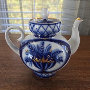 Könnte beinhalten: Eine weiße Porzellan-Teekanne mit blauen floralen und geometrischen Mustern, akzentuiert mit goldenen Verzierungen. Die Teekanne hat einen gebogenen Griff, einen Ausguss mit goldener Spitze und einen Deckel mit kleinem Knauf. Klassisches, elegantes Design.