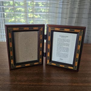 May include: A double picture frame in dark brown wood, with gold and orange inlaid detailing. The frame is open, showing a blank space on one side and a printed document on the other. Hinged design.